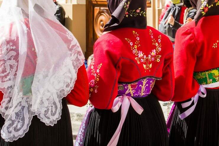 Donne di spalle vestite con i tipici abiti sardi, in processione. Velo bianco di pizzo per una e altre due ragazze con giacca rossa, cintura viola o verde, e gonna nera 